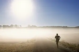 Roads: Man training running on a gravel road in the morning fog in a rural landscape with the sun shining through the fog