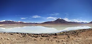 Volcano: Wide view of a volcano with a reflecting lake and rocky shore under a blue sky